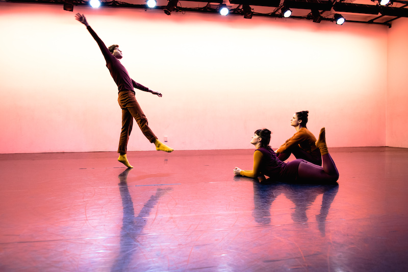 Two female dancers sit on the floor and look up at a male dancer perform. He balances on one leg as his right leg dangles in the air and his right arm extends to the ceiling